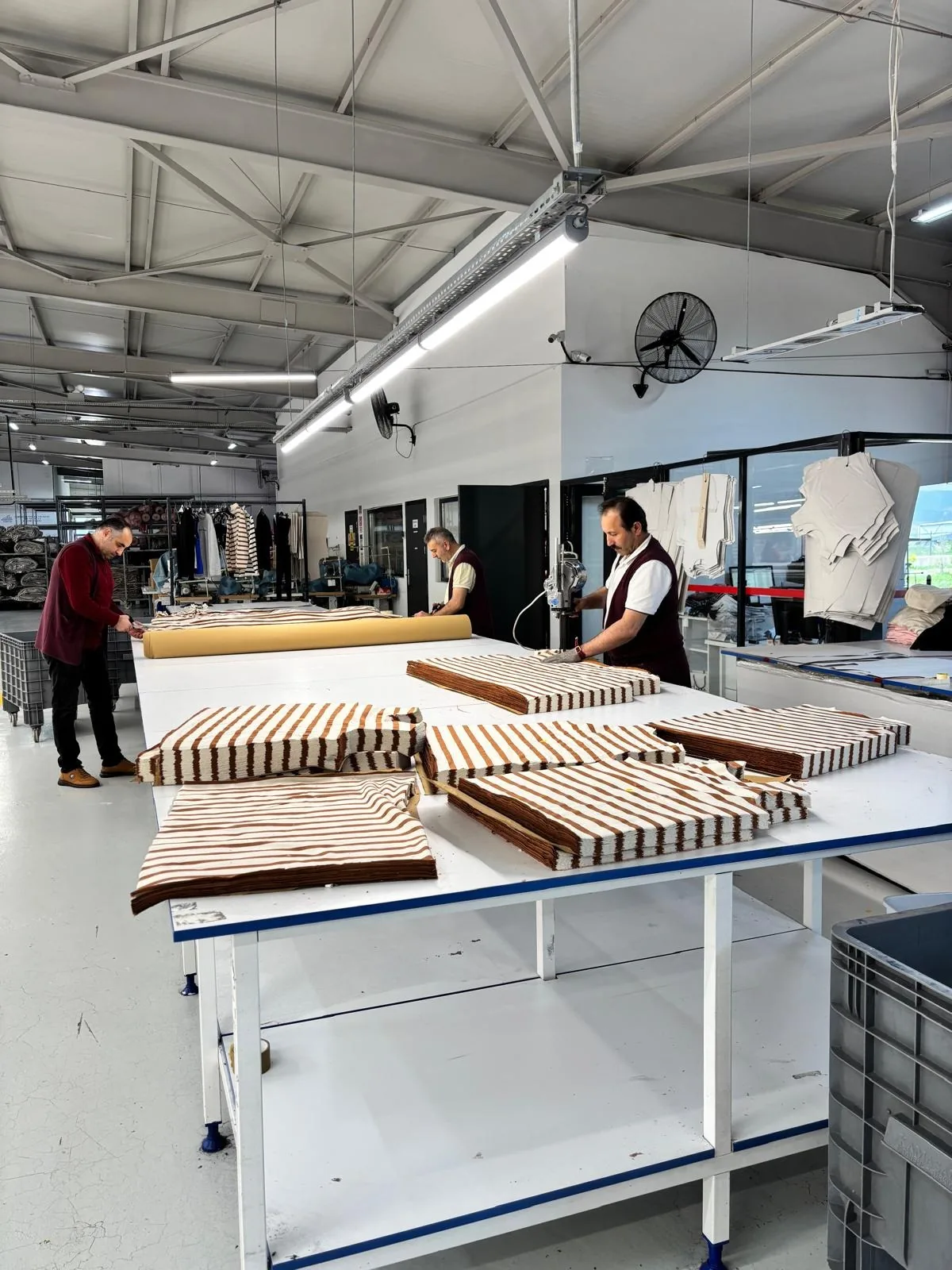 Erbaa fabrikasında son kontrol masasında üst üste dizilmiş çizgili örme parçalar; arkada salonda çalışan personel